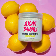 Load image into Gallery viewer, A clear jar filled with whipped body butter is laying on six lemons on a baby pink background. It has a white label with The Sugar Bandits logo written in hot pink in the center, below that whipped body butter written in black, lemon sorbet written in yellow and 3.6oz/102g written in black centered below that.