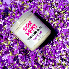 Load image into Gallery viewer, A clear jar filled with whipped body butter is laying askew on a purple background covered in royal purple flowers. It has a white label with The Sugar Bandits logo written in hot pink in the center, whipped body butter written in black, black flower written in purple and 3.6oz/102g written in black centered below that.