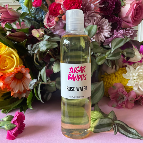 A clear bottle of rose water is sitting in front of baby blue hydrangeas, pink, white, and yellow roses, lilac, pink, and orange carnations, light pink lilies and tiger lilies and light green and green leaves on a baby pink surface. The bottle has a white label with The Sugar Bandits logo written in hot pink in the center and rose water and Net wt. 8.5oz/240 g written in black centered below that.