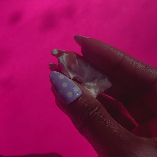 A hand with long nails painted with daisies is rubbing creamy whipped body butter back and forth across their fingers on a pink background.