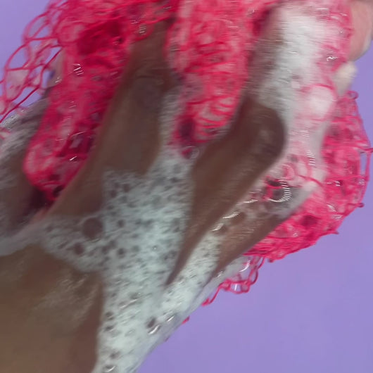 A video of a pink sudsy exfoliating sponge scrubbing an arm in front of a lilac background.