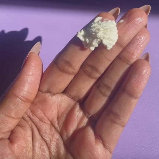 A hand with long nude nails is rubbing creamy whipped body butter back and forth across their fingers on a pink background.