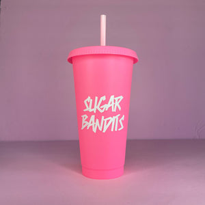 A hot pink plastic cup with a lid and straw that looks like a fast food cup is on a baby pink background. It has the The Sugar Bandits logo in white written in the center of the cup.