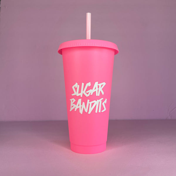 A hot pink plastic cup with a lid and straw that looks like a fast food cup is on a baby pink background. It has the The Sugar Bandits logo in white written in the center of the cup.