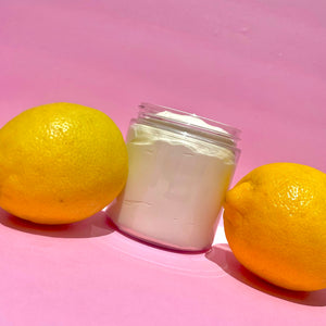 A clear jar filled with whipped body butter with a lemon on either side against a pink background.