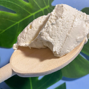 A small wooden spoon with whipped body butter on it takes up most of the frame. It is on a baby blue background with Monstera leaves, and shows the airy bubbles in the whipped body butter.