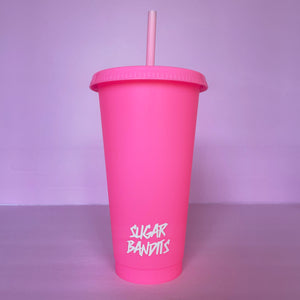 A hot pink plastic cup with a lid and straw that looks like a fast food cup is on a baby pink background. It has the The Sugar Bandits logo written in white at the bottom of the cup.