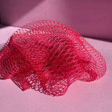 Load image into Gallery viewer, A pink exfoliating sponge is sitting on a baby pink background. You can see the netted texture and it is illuminated by sunlight streaming in from a window you can’t see.
