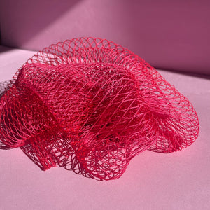 A pink exfoliating sponge is sitting on a baby pink background. You can see the netted texture and it is illuminated by sunlight streaming in from a window you can’t see.