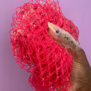 A pink sudsy exfoliating sponge is held in a hand with the thumb facing outward. It has a long nude nail with a purple heart painted at the base and it is in front of a baby pink background. 