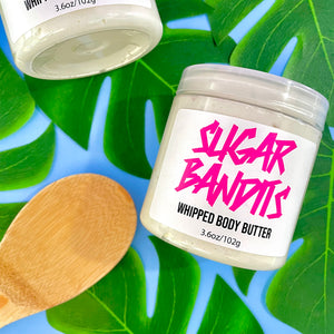 Two clear 3.6 oz jars, filled with whipped body butter, on a baby blue background covered with Monstera leaves. One is out of frame in the top left and the other is in frame in the center right, there is a medium wooden spoon off center in the bottom left corner. The jar in frame has a white label with The Sugar Bandits logo written in hot pink in the center and whipped body butter and 3.6oz/102g written in black centered below that.